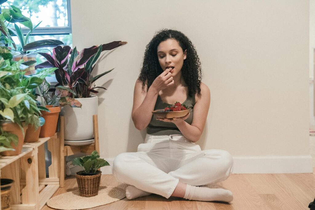 A woman with curly hair sits on the floor eating fresh berries, surrounded by potted plants.