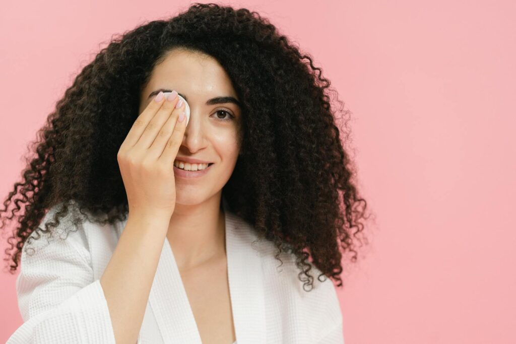 Young woman with curly hair using a cosmetic pad for skincare on a pink background.