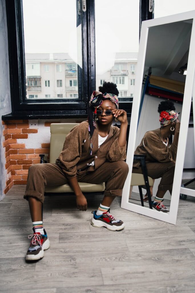 Stylish black woman in trendy outfit posing by a window with a mirror reflection.