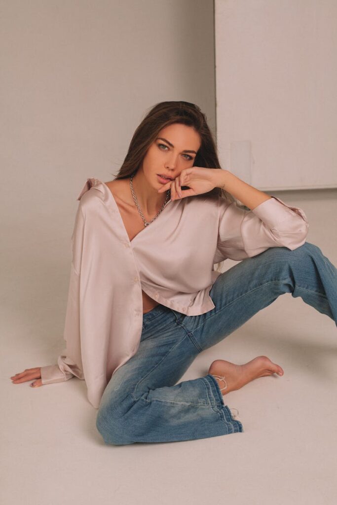 Woman with long brown hair sitting on floor in casual blouse and jeans, looking pensive.