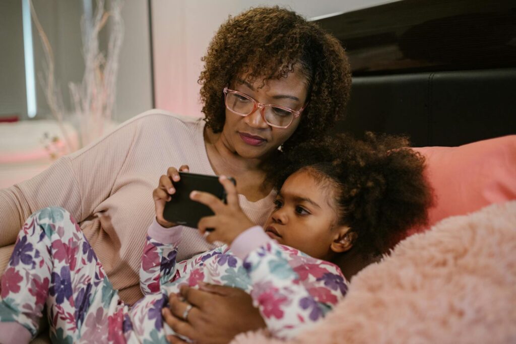 A mother and daughter enjoy bedtime together, relaxing with a smartphone in a cozy bedroom setting.