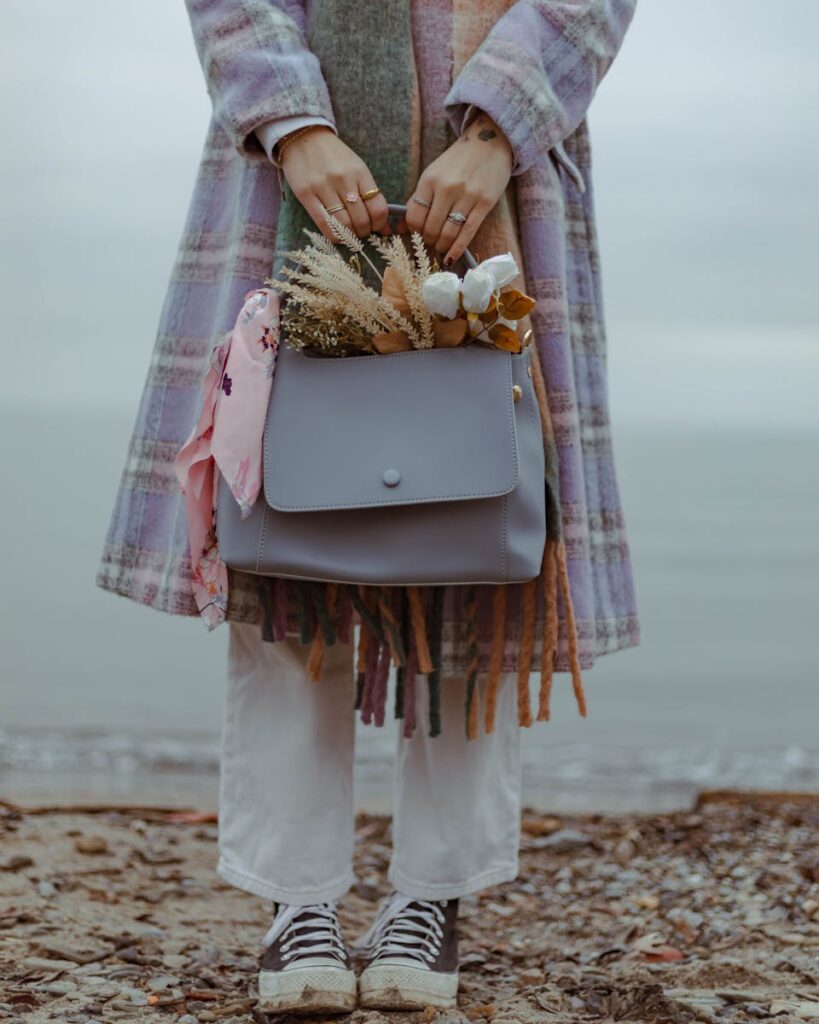 A woman in a coat holds a handbag with flowers, standing by the beach.