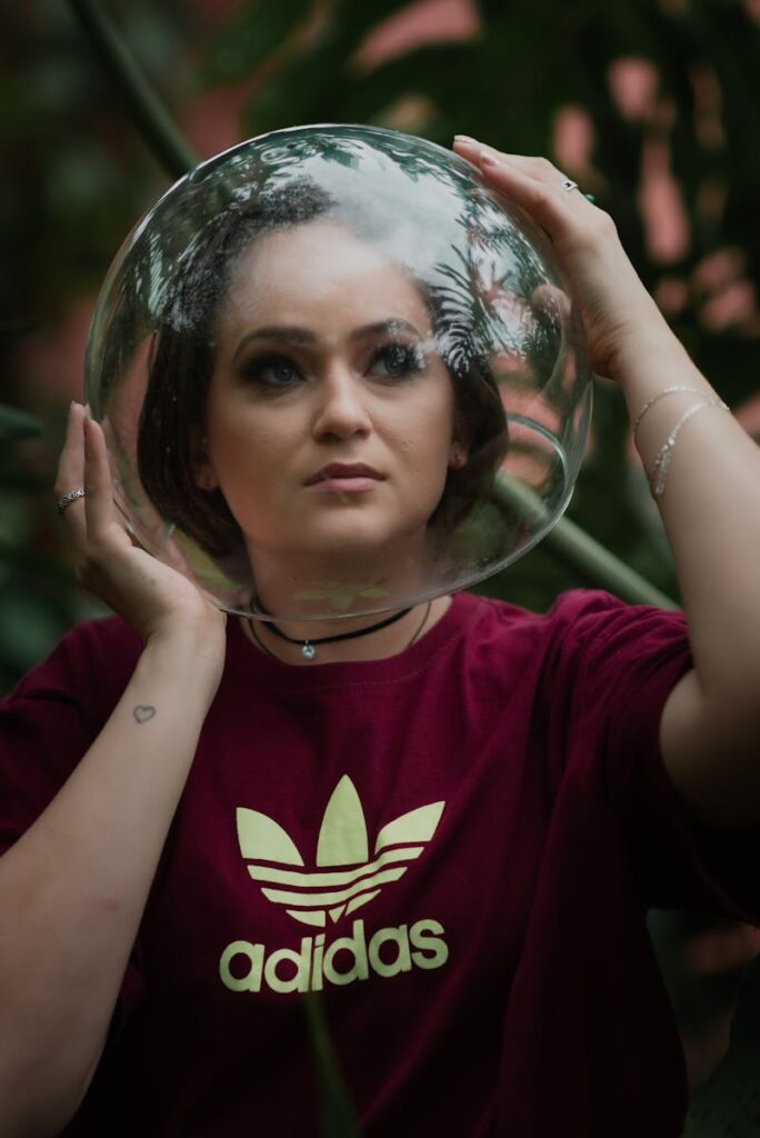 A woman in a burgundy adidas shirt poses creatively with a glass headpiece in an outdoor setting.