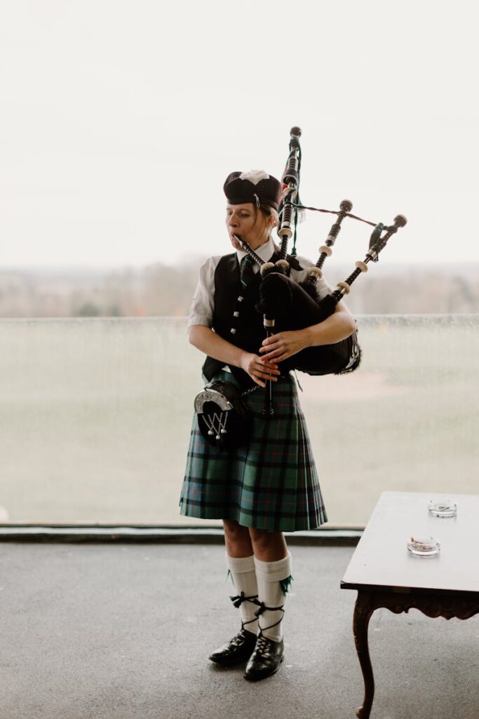 A woman playing bagpipes in traditional Scottish attire, indoors.