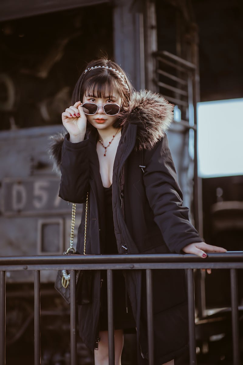 Elegant woman in sunglasses and fur jacket poses by railing outdoors, conveying fashion and confidence.
