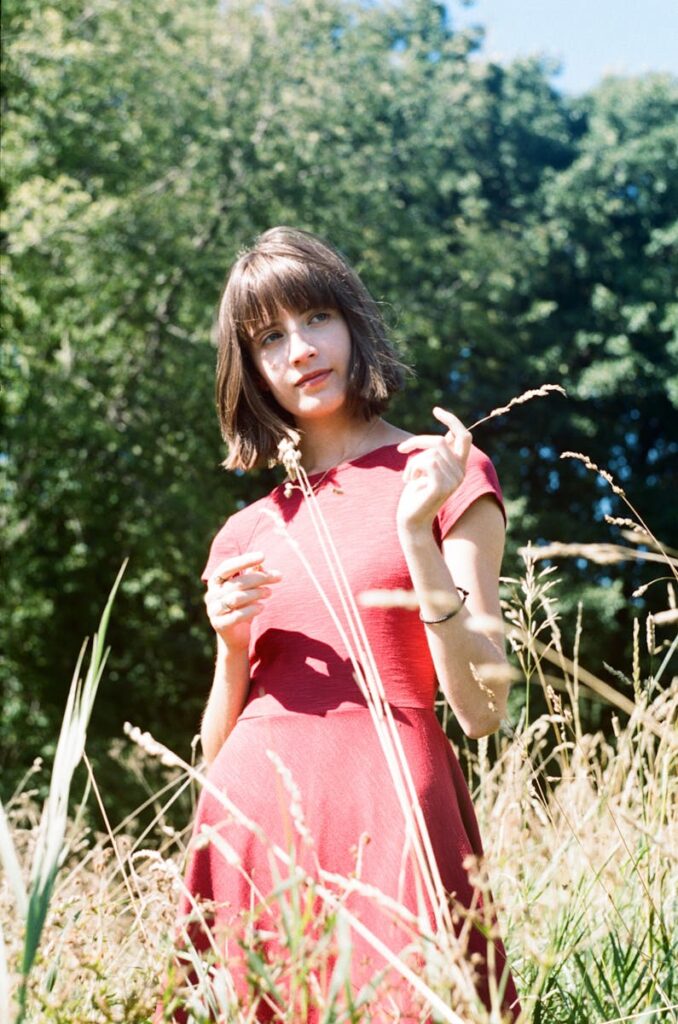 Woman with brown hair in a red dress standing in a sunlit meadow surrounded by greenery.