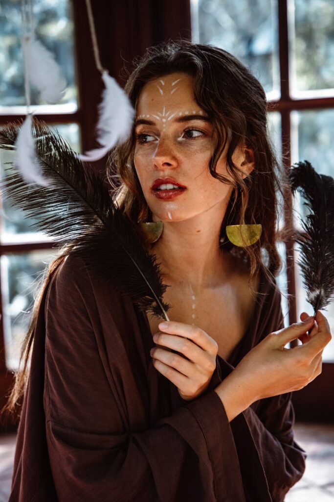 Creative portrait of a woman with face paint holding feathers indoors, captured in natural light.