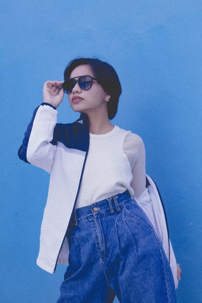 Fashionable woman posing in denim jeans and sunglasses with a vibrant blue backdrop.