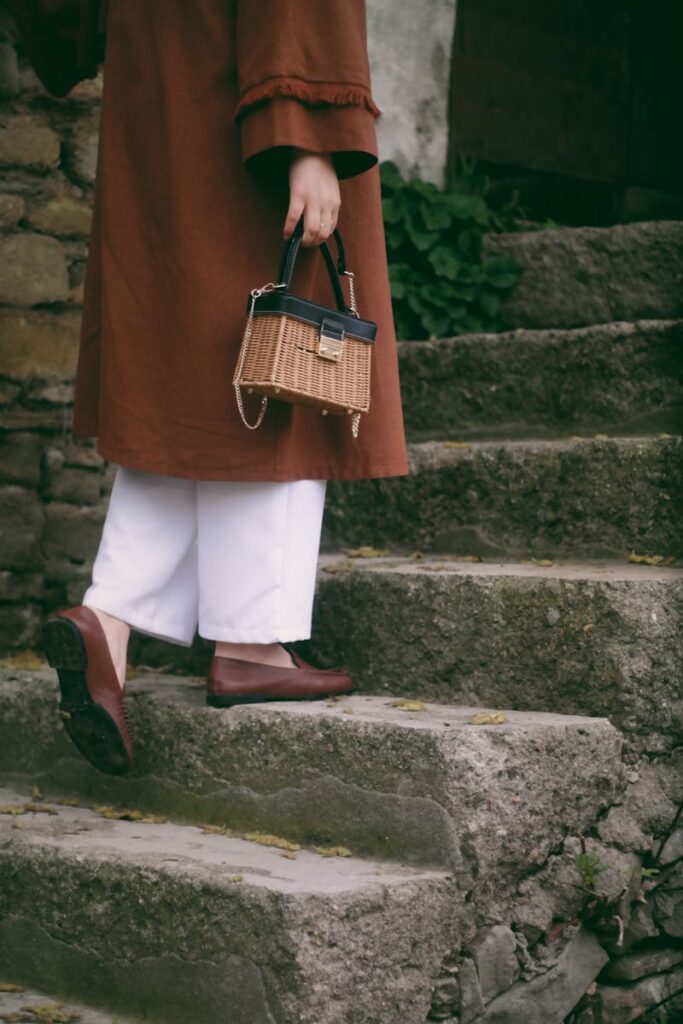 Woman in brown coat with a wicker bag walking up stone steps outdoors.