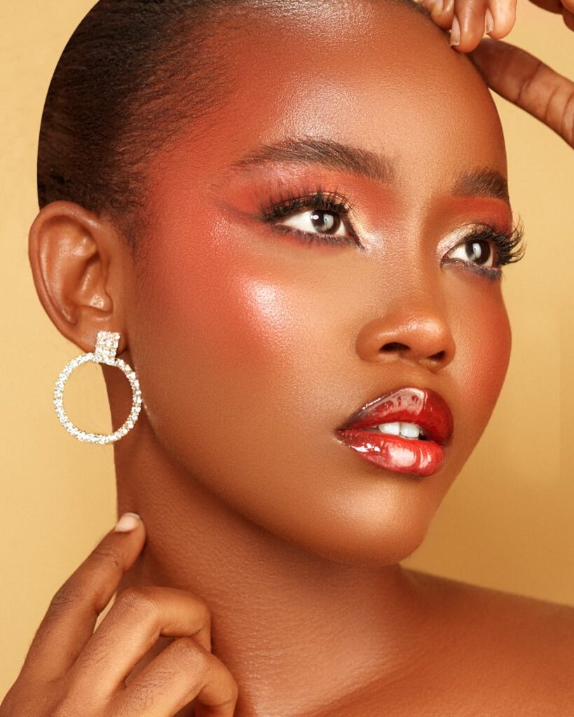 Close-up portrait of a model with glowing makeup and statement earrings on yellow backdrop.
