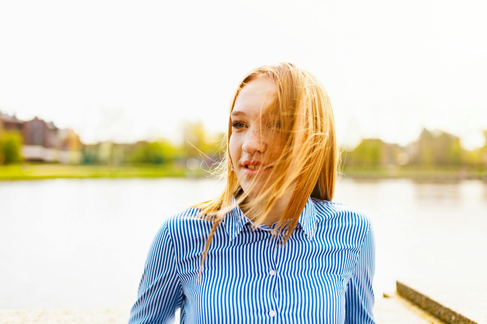 Portrait of a young woman with blonde hair smiling by a lake on a sunny day.