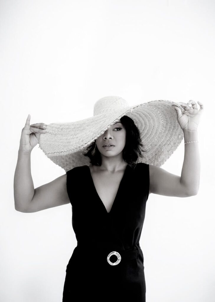 Elegant woman posing in a black outfit with a large straw hat in a studio setting.