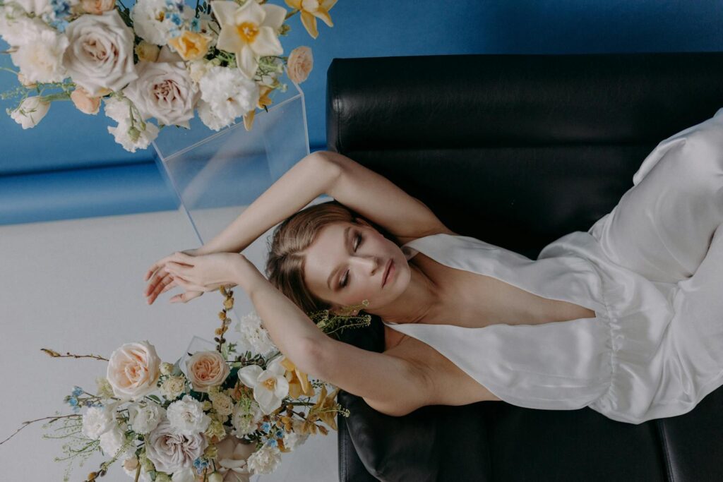 A woman in a stylish white jumpsuit relaxes gracefully with closed eyes, surrounded by elegant floral arrangements.