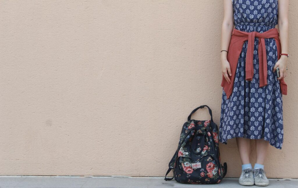 Stylish woman in a patterned dress and sneakers standing with a floral backpack against a neutral wall.