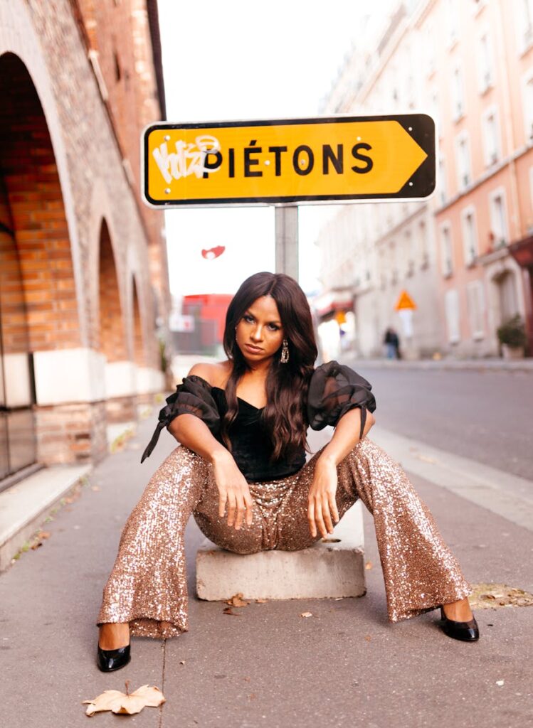 A fashionable woman sitting on a city street in bold sequin pants and a pedestrian sign.