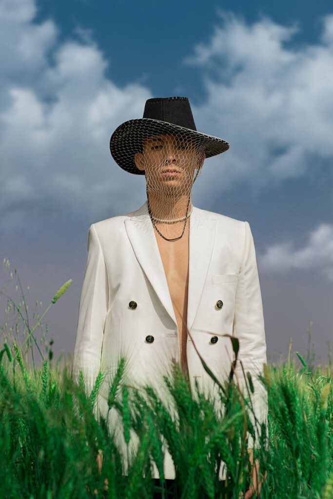 A fashionable man in a white blazer and hat stands in a wheat field under a vivid sky.
