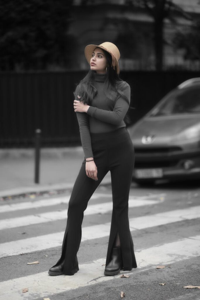 Fashionable woman poses confidently on city street, wearing trendy attire with a hat and flared pants.