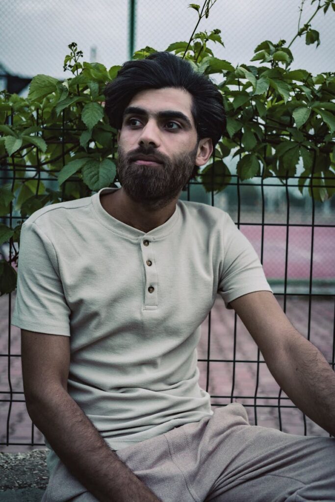 A bearded man in a crew neck t-shirt sits seriously beside a metal fence with climbing plants.