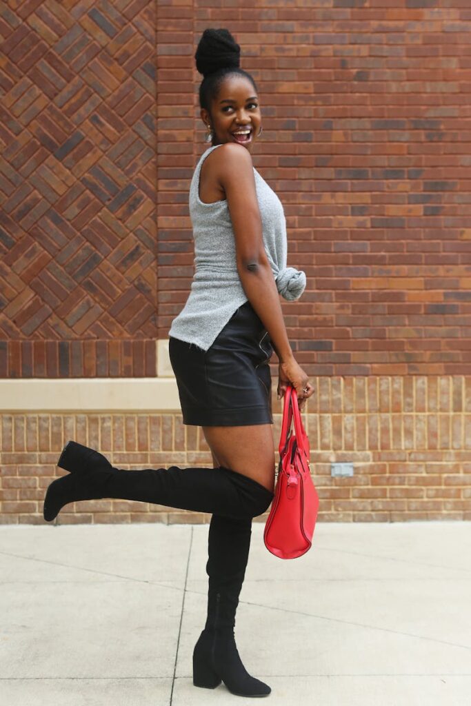 Chic woman poses happily in a modern outfit with a red bag against a brick wall.