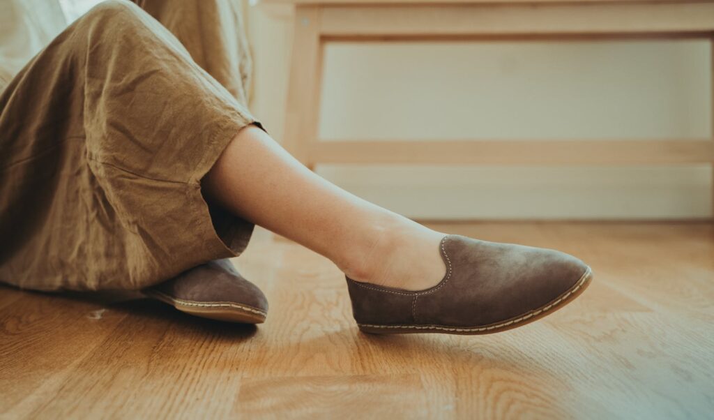 Close-up of a woman's legs wearing brown flats on a wooden floor, captured indoors.