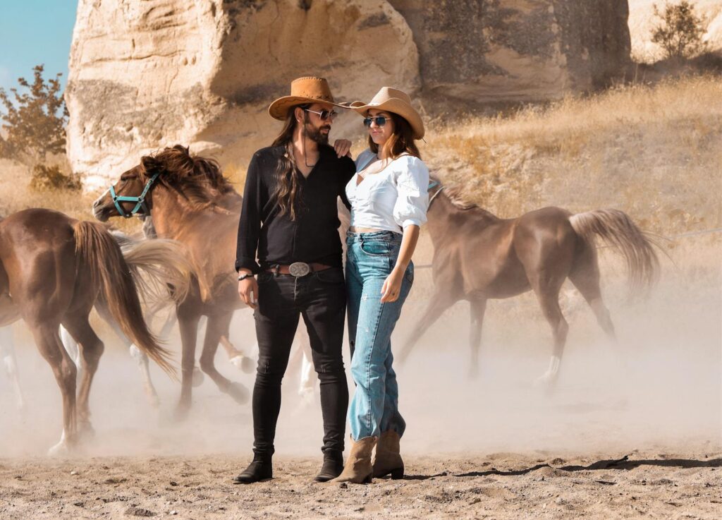A fashionable couple in cowboy hats stands confidently as horses roam in a rustic outdoor setting.