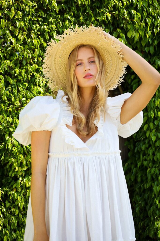 A fashionable woman in a white dress and straw hat poses outdoors in a sunny, vibrant setting.