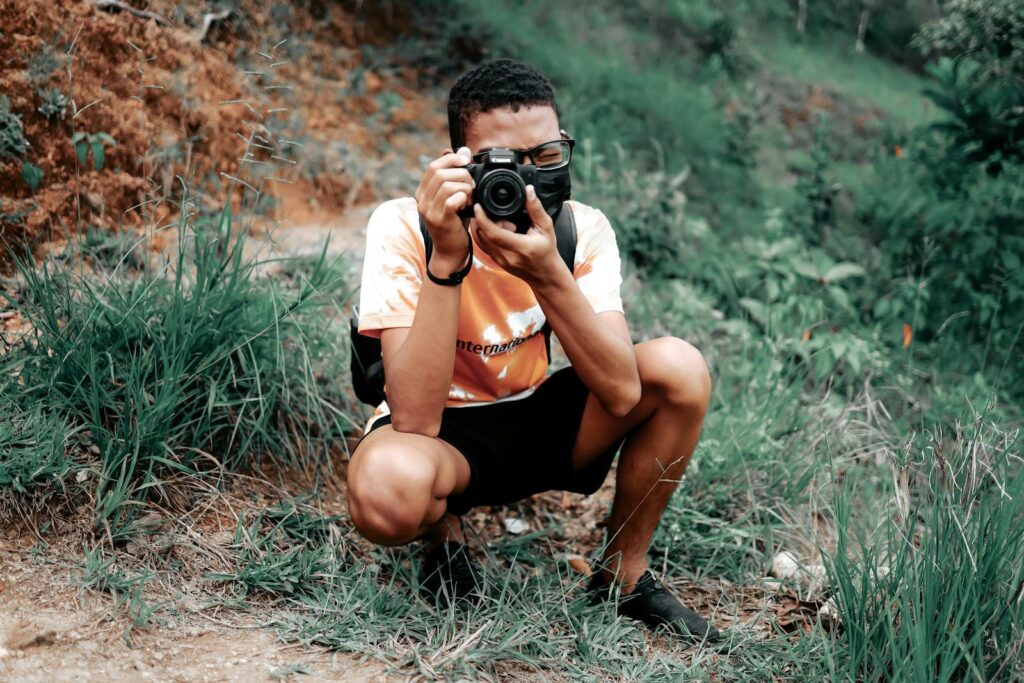 A young man crouching outdoors captures nature through his camera lens.