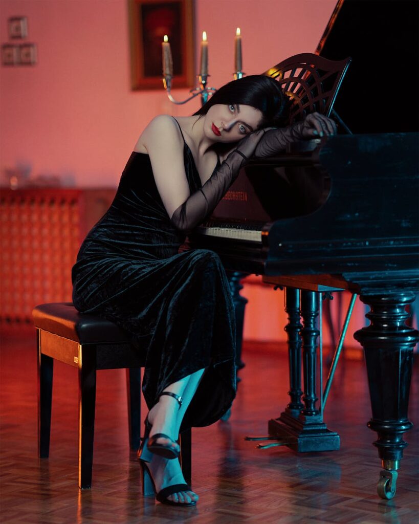 A beautifully dressed woman in formal evening wear sitting by a piano in a dimly lit room.