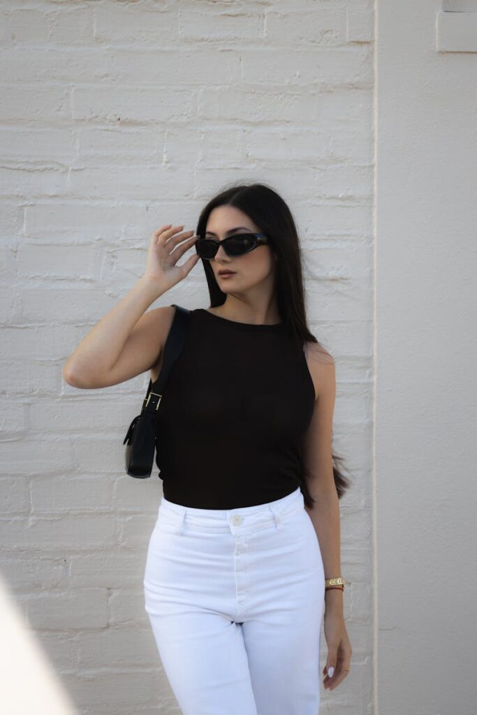 Fashionable woman wearing sunglasses and chic outfit posing outdoors against a white brick wall.