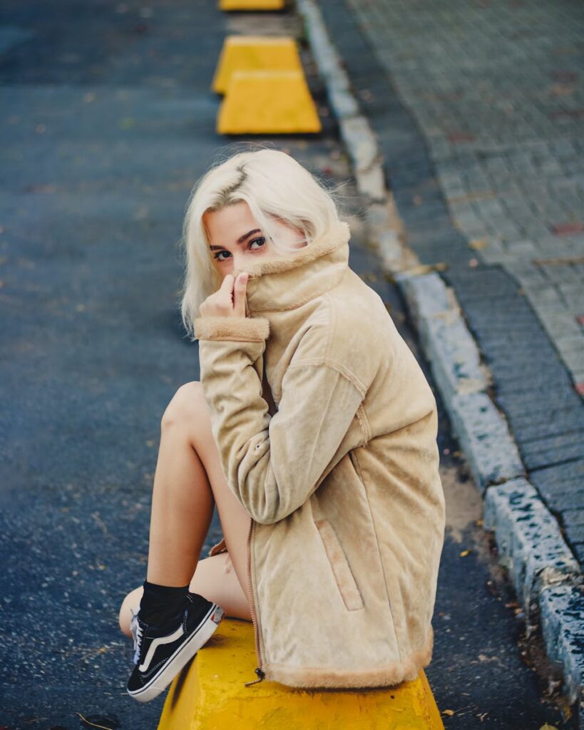 Blonde woman sitting on a curb wearing a suede jacket and black sneakers, exuding stylish vibes.