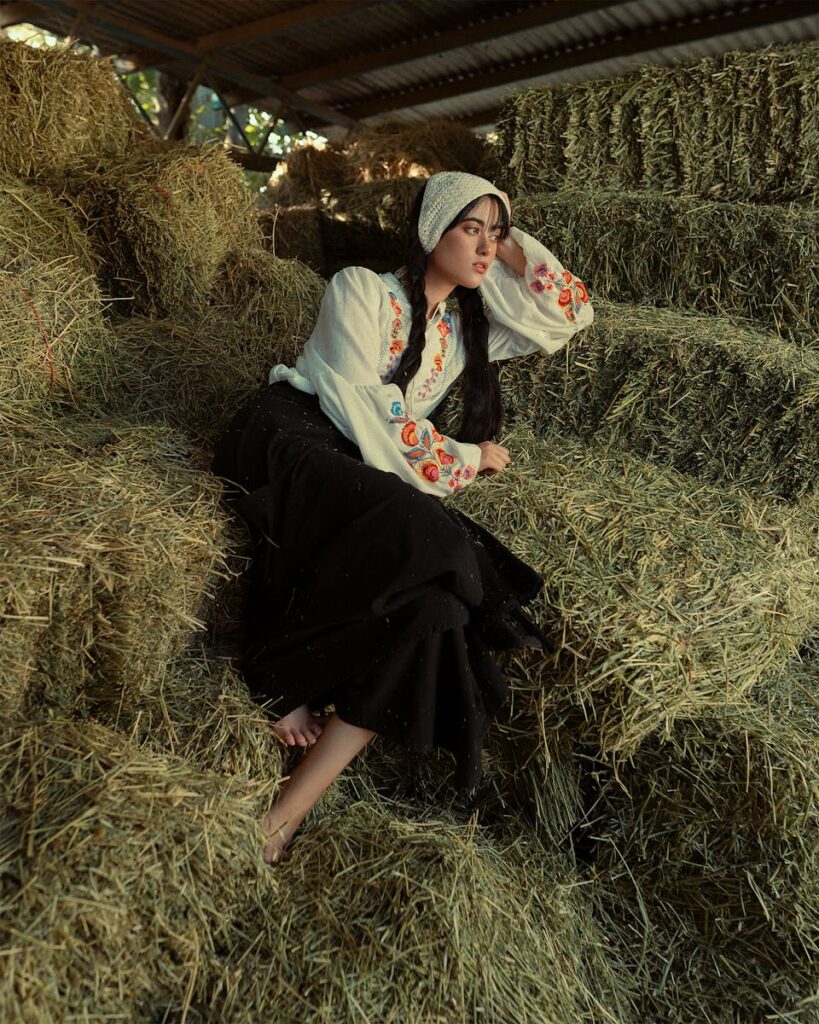 Woman in traditional attire resting on hay in a serene farm setting.