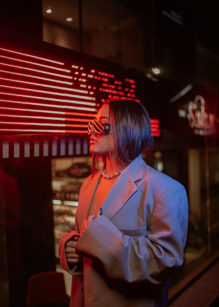 Fashionable woman in sunglasses and oversized blazer posing under neon lights at night.