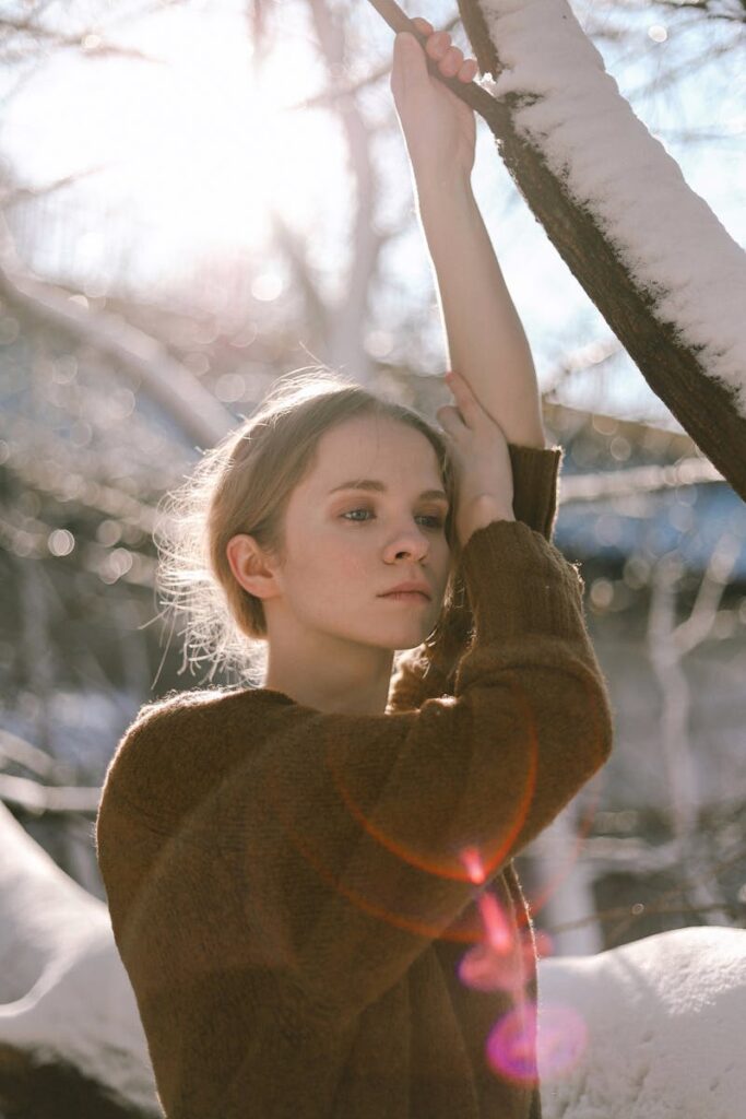 A young woman poses outdoors with sunlight streaming through snowy branches.