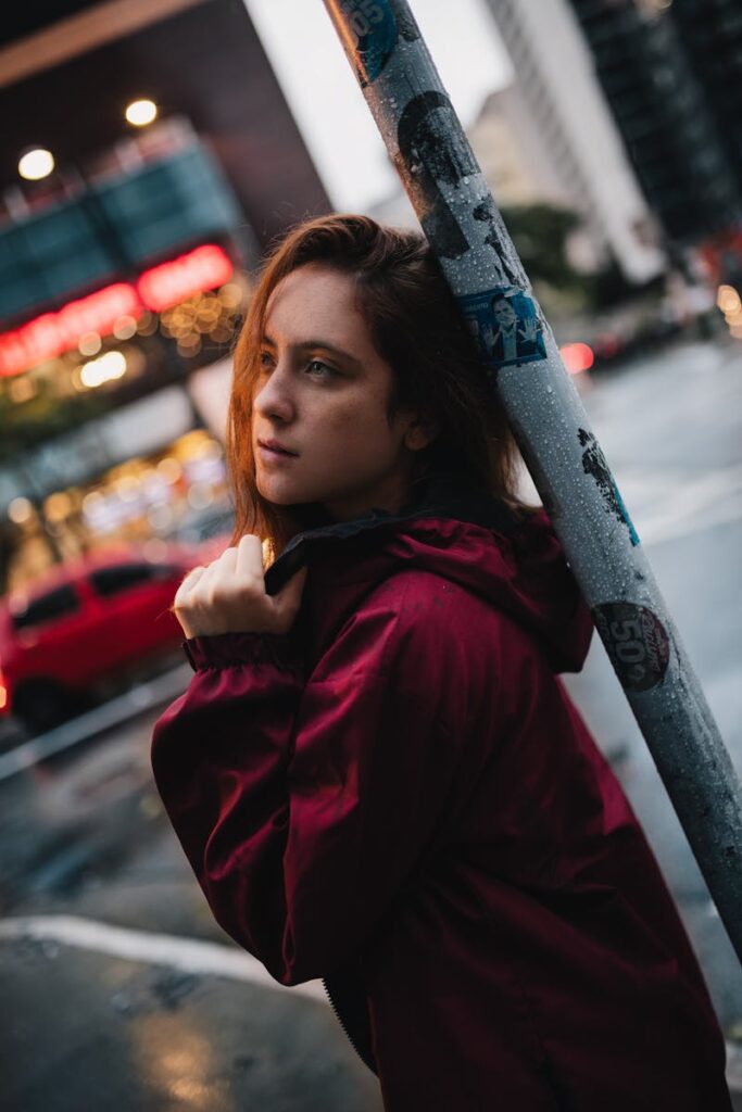 Young woman in a red jacket leans on a post in a rainy city street.