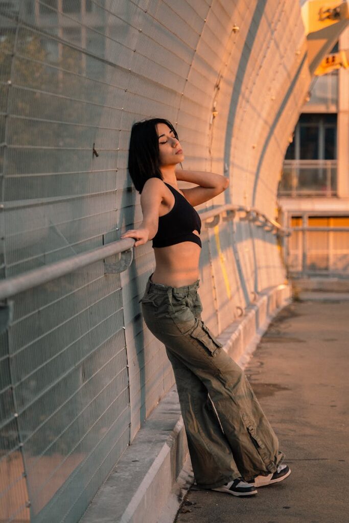 Stylish woman in cargo pants and black top posing on an urban bridge at sunset.