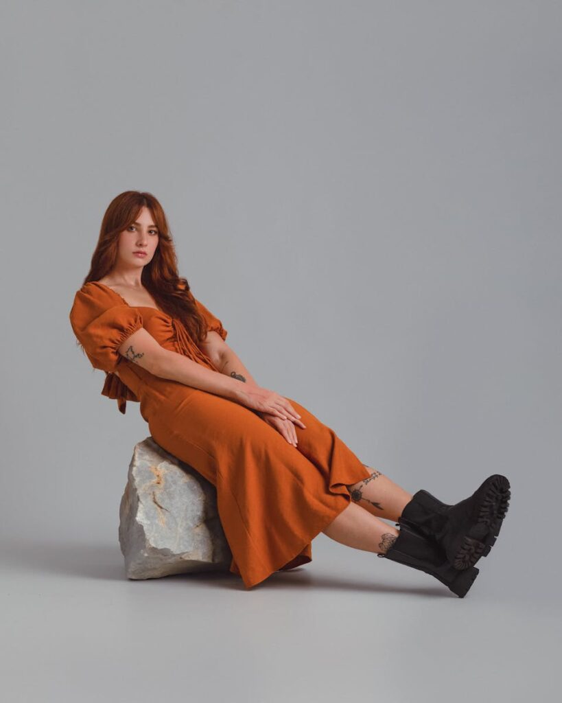 Fashion portrait of a woman in an orange dress and boots posing indoors on a rock.