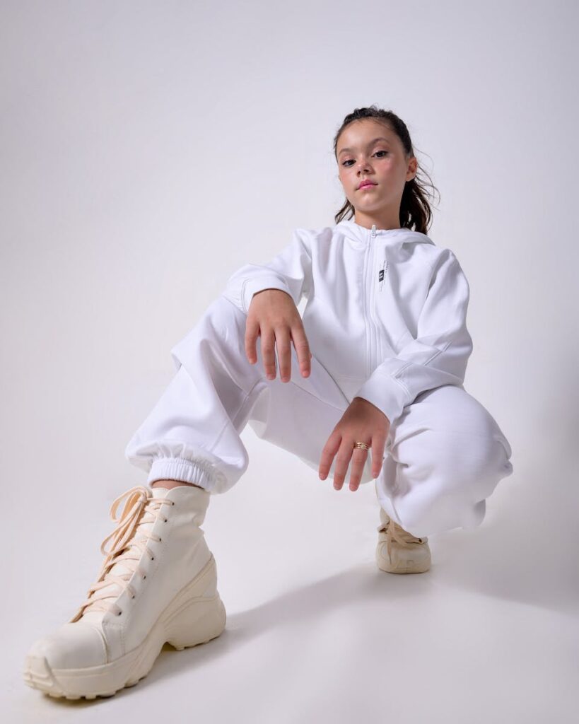 Fashionable young girl posing confidently in a white tracksuit and boots against a gray background.