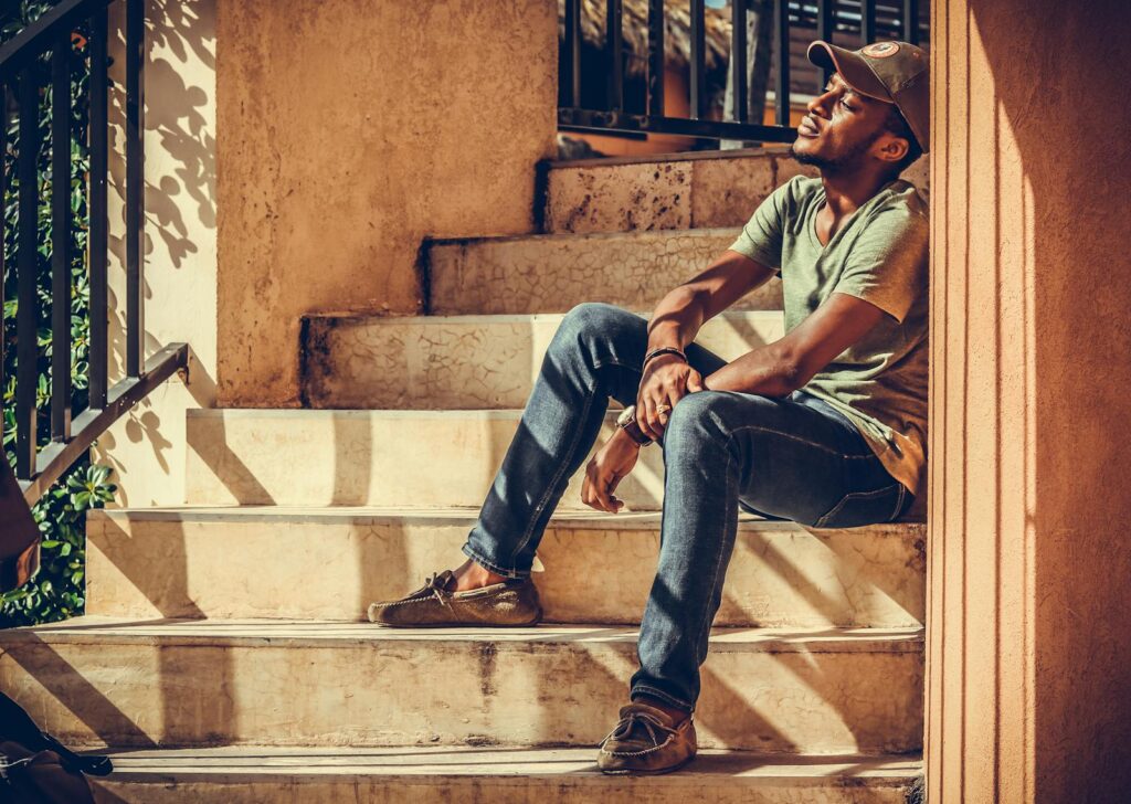 A fashionable man in casual attire enjoys sunlight on an outdoor staircase, exuding a relaxed vibe.