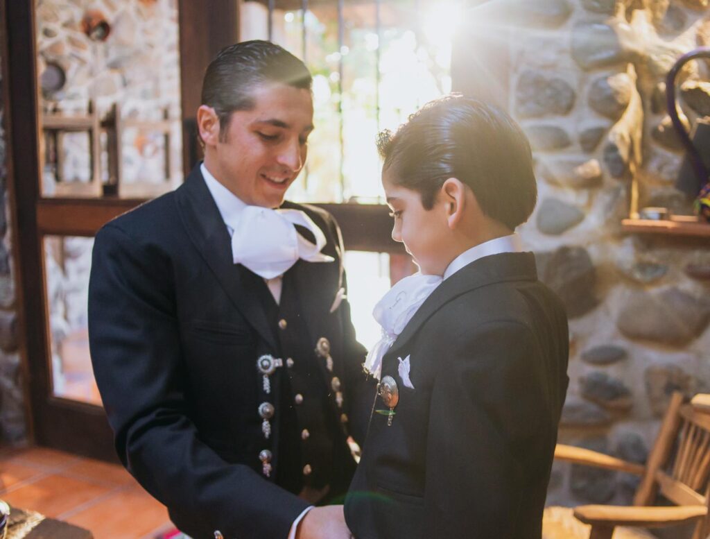 A father and son in traditional Mexican suits share a tender moment indoors.