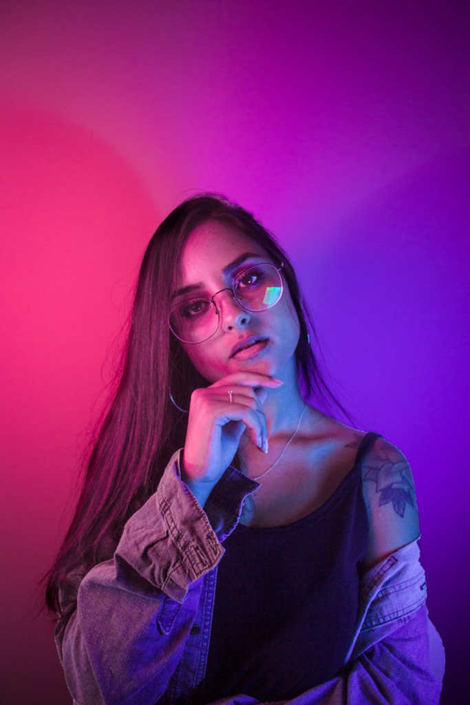Stylish woman in neon lighting, wearing glasses and denim, posing confidently.