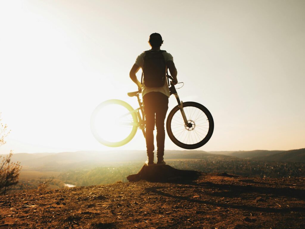 A cyclist stands silhouetted with a mountain bike on a hilltop during sunset, evoking adventure and freedom.