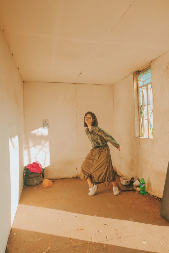 Young woman in a stylish outfit posing in a sunlit minimalistic room with rustic decor.