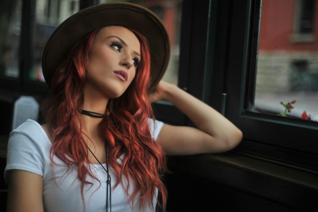 Portrait of a stylish redhead woman looking out a window indoors, wearing a hat.