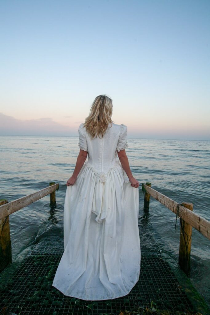 Woman in a white dress standing on a pier facing a serene sea at sunset.