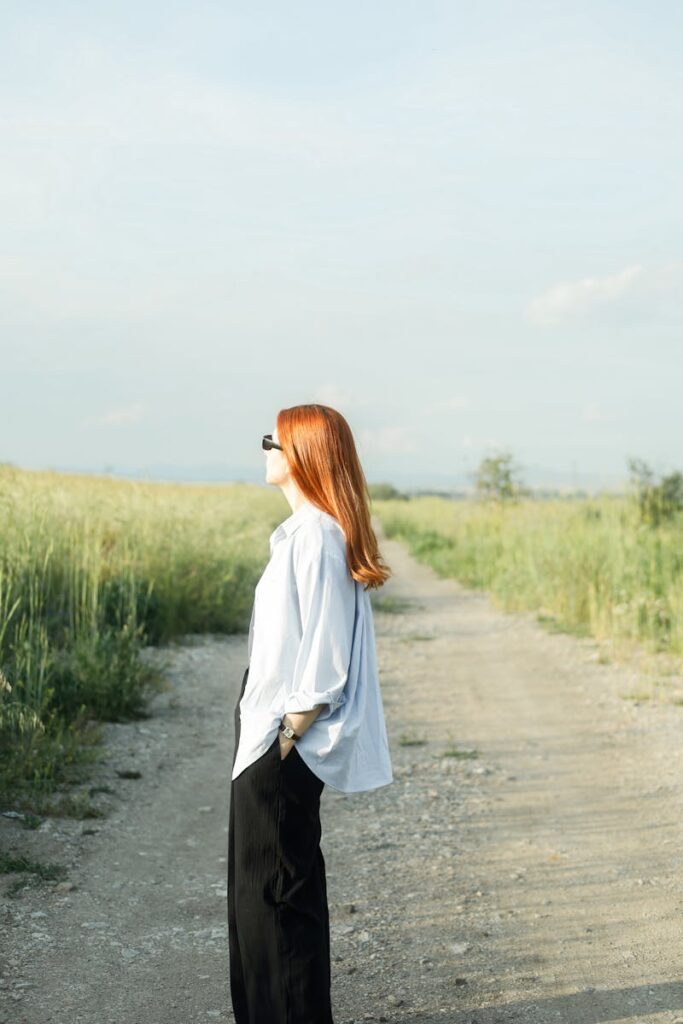 Redhead woman standing on a rural dirt road, side view in sunlight.