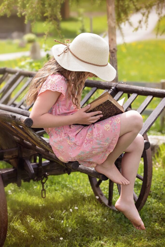 A young girl in a summer dress reads a book on a rustic cart in a sunny garden.