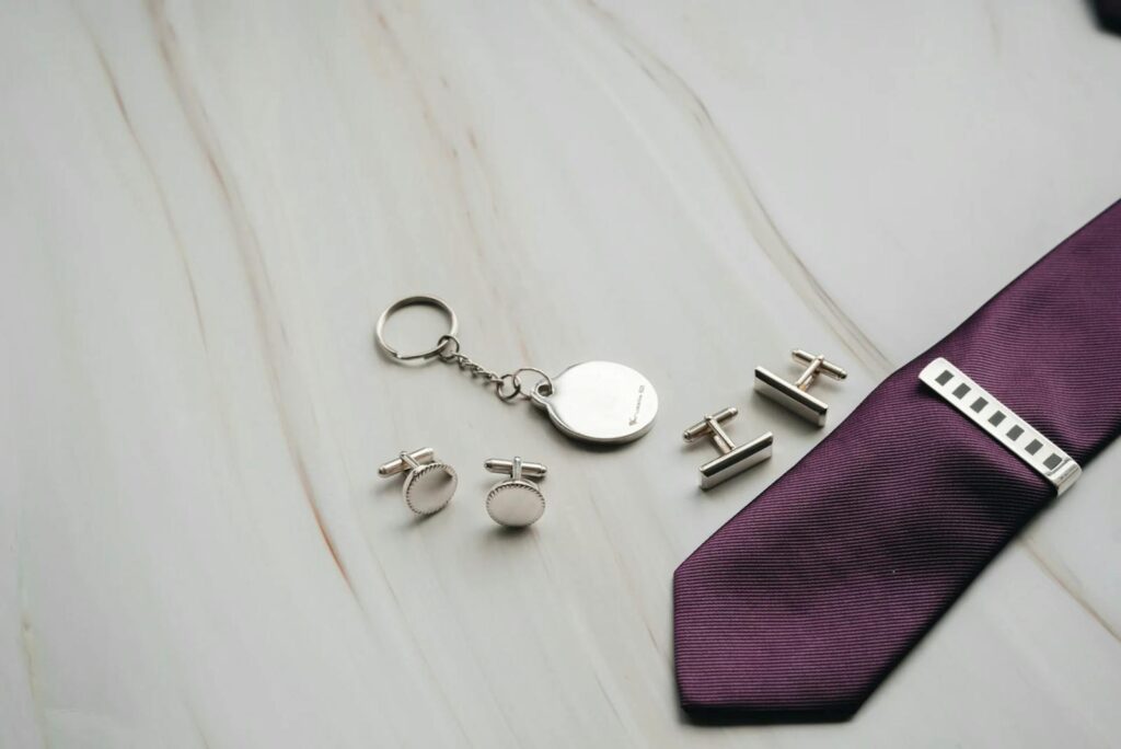 Flat lay of luxury men's accessories with a purple tie, cufflinks, and keychain on a marble surface.