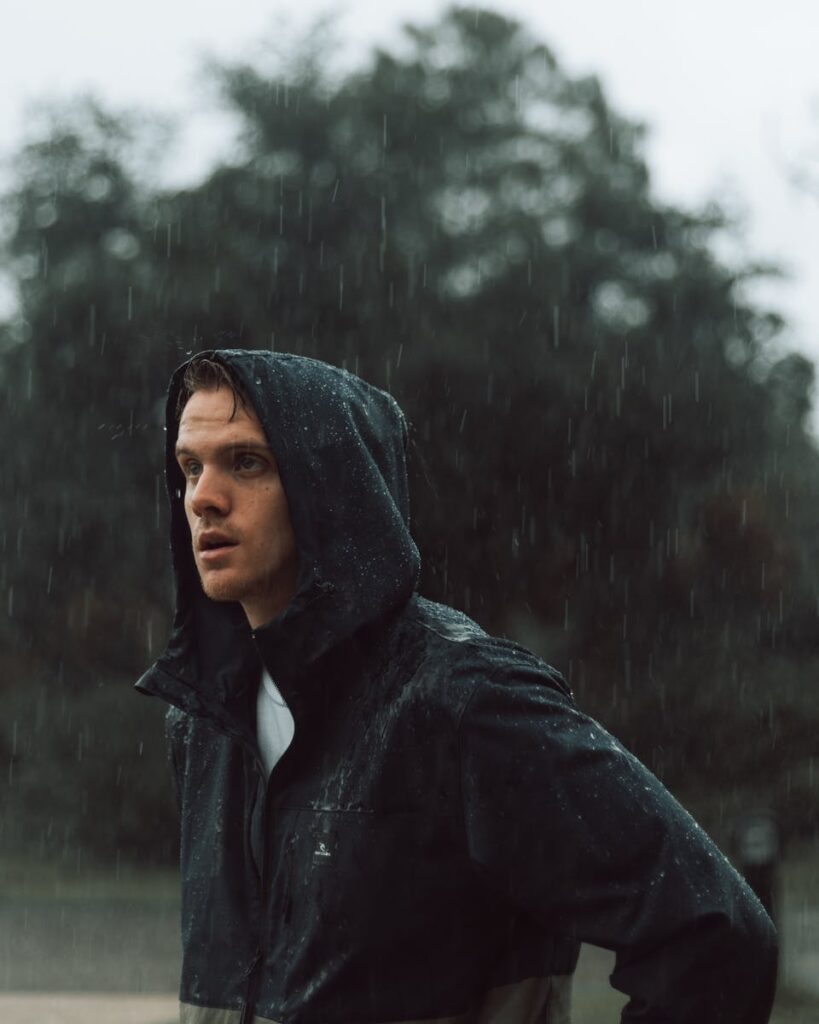 Moody portrait of a man standing outdoors in a rainy setting, wearing a hooded jacket.