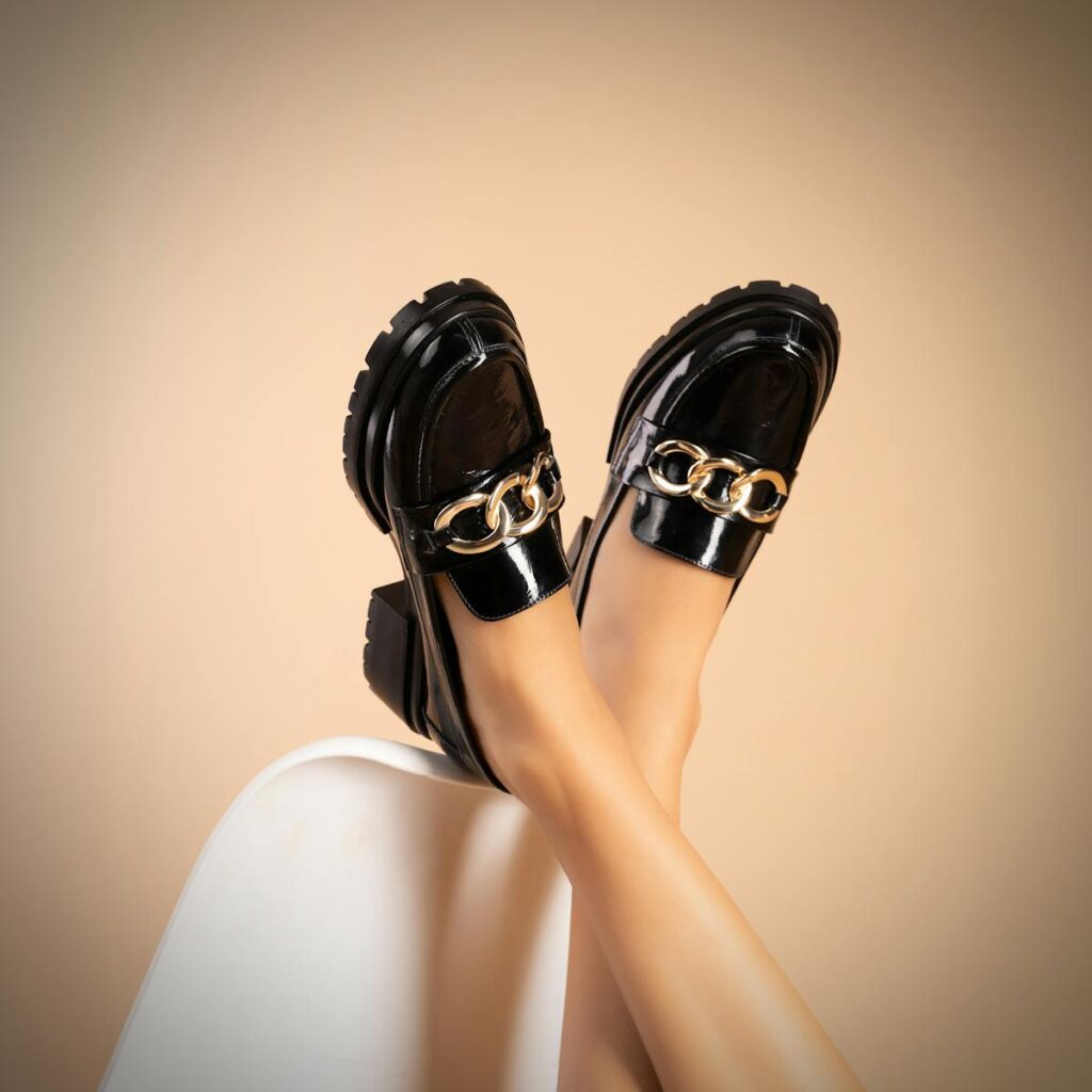 Close-up shot of fashionable black patent leather loafers with gold chain detail against a beige backdrop.