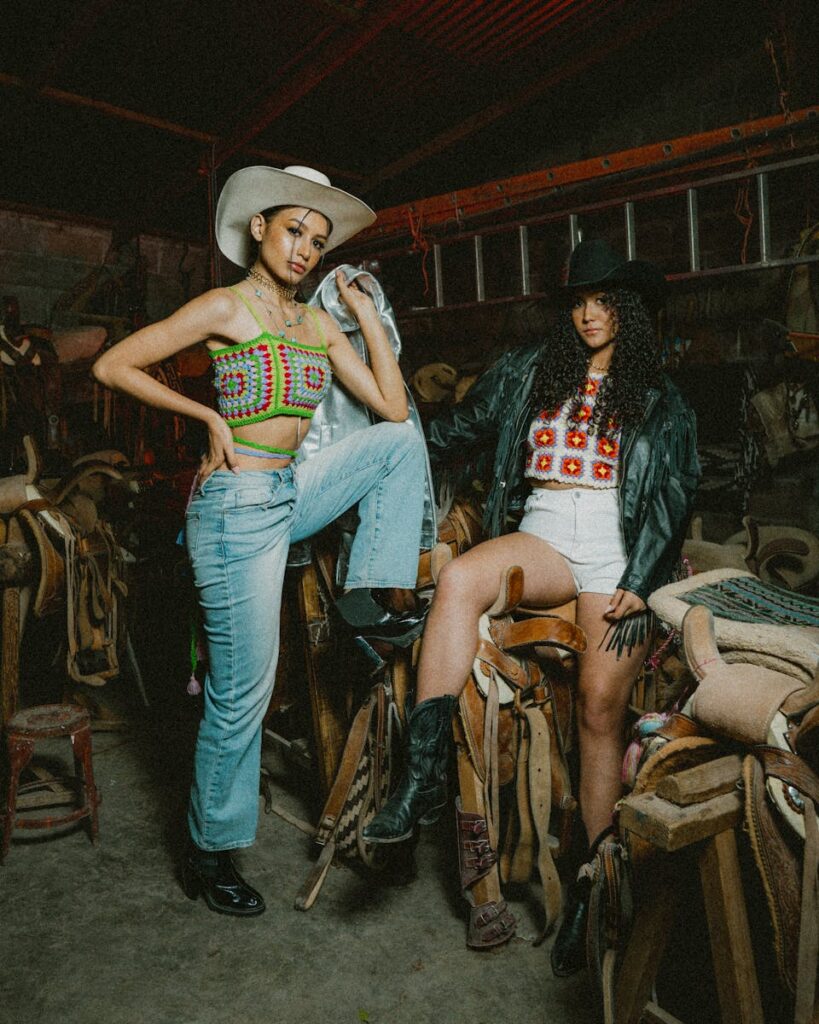 Two young women in cowboy attire pose in a rustic setting for a fashion shoot.